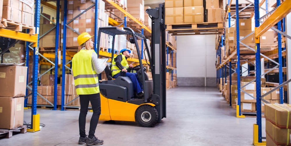 image of someone within a warehouse holding a clipboard whilst the other is on an orange forklift, proceeding to lift an object from the rack.